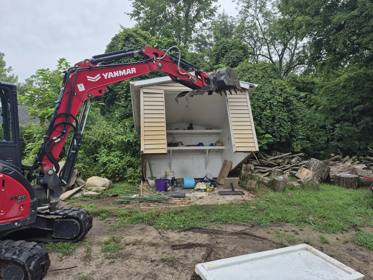 Mini excavator lifting a small wood shed off its base during a teardown