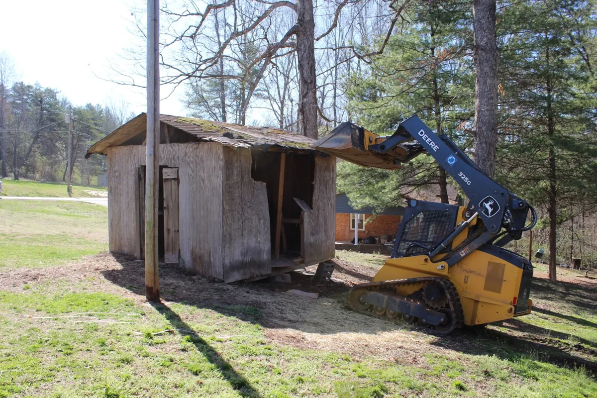 Compact track loader pushing over an old wooden shed in a yard